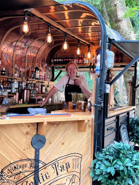 A bartender smiles from behind a wooden bar in a forested outdoor setting.