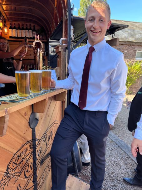Man in a white shirt and red tie, standing by a bar with glasses of beer.
